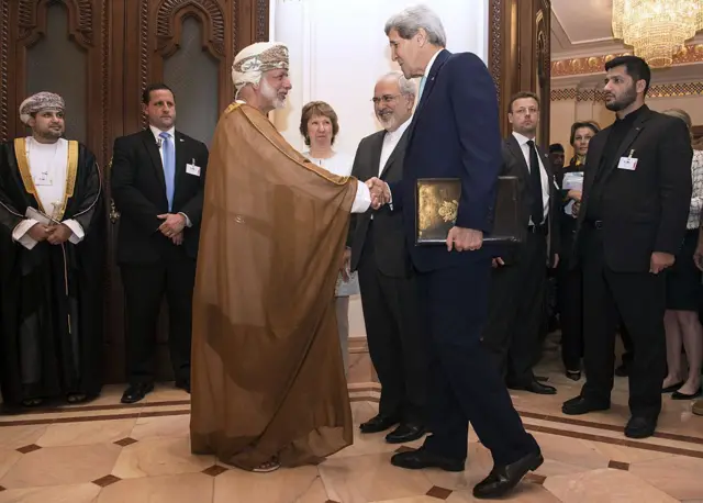 وزير الخارجية الأمريكي آنذاك جون كيري (يمين) مرتدياً بدلة رسمية يصافح وزير الخارجية العماني يوسف بن علوي الذي يرتدي زي بلاده الرسمي، في مسقط في 9 نوفمبر/تشرين الثاني 2014، وكان من بين الحاضرين في الاجتماع أيضا مسؤولة السياسة الخارجية السابقة في الاتحاد الأوروبي كاثرين أشتون ووزير الخارجية الإيراني محمد جواد ظريف.