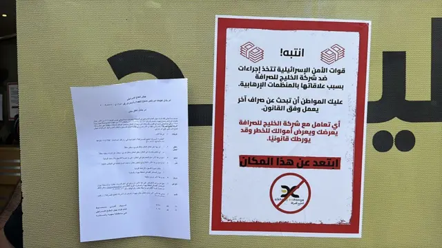 عُلّقت ملصقات تحذيرية على نوافذ المكاتب، تحذر المواطنين من إجراء أي معاملات مالية، بعد أن نفذت القوات الإسرائيلية مداهمات متزامنة لفروع شركة "الخليج" للصرافة في رام الله، الضفة الغربية، في ٢٧ مايو/أيار ٢٠٢٥.