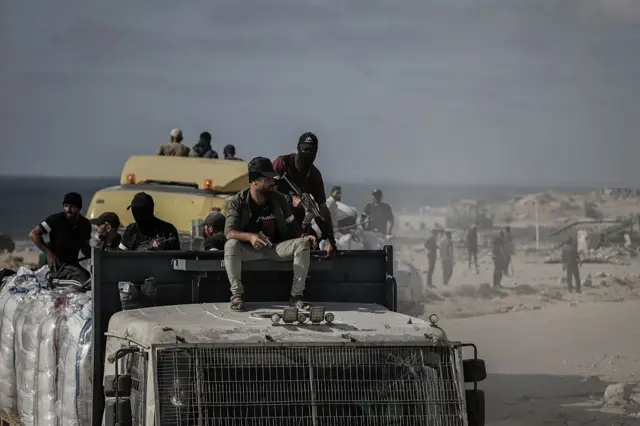 شاحنة يعتليها مجموعة من الأشخاص إضافة إلى بضائع مساعدات في الخلفية أرض ترابية. الصورة التقطت نهاراً.