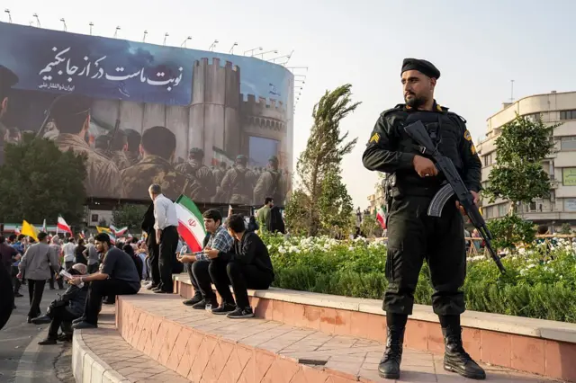 ضابط شرطة إيراني يقف، بينما يلوح المتظاهرون بالأعلام ويهتفون خلال تجمع لتكريم القوات العسكرية الإيرانية بالعاصمة طهران، عقب إعلان وقف إطلاق النار بين إسرائيل وإيران في ٢٤ يونيو/حزيران ٢٠٢٥.