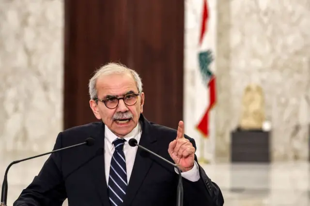  رئيس الحكومة اللبنانية، نواف سلام واقفاً وأمامه ميكرفون، مرتدياً سترة سوداء وقميصاً أبيض  وربطة عنق يمتزج لونها ما بين الأبيض والأسود.