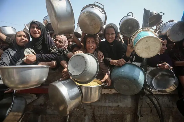 فلسطينيون جائعون، بينهم نساء وأطفال، يحملون أواني الطعام، ينتظرون الحصول على الطعام