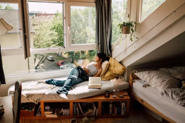 شابة تسترخي على مقعد النافذة، محاطة بغرفة نوم مريحة وهادئة مع إضاءة طبيعية ونباتات.