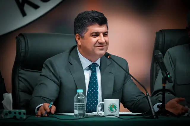 لاهور شيخ جنكي يجلس على مكتب وأمامه مكيروفون، مرتدياً سترة رمادية اللون وربطة عنق زرقاء دوائر صغيرة بيضاء، وقميصاً أبيض.