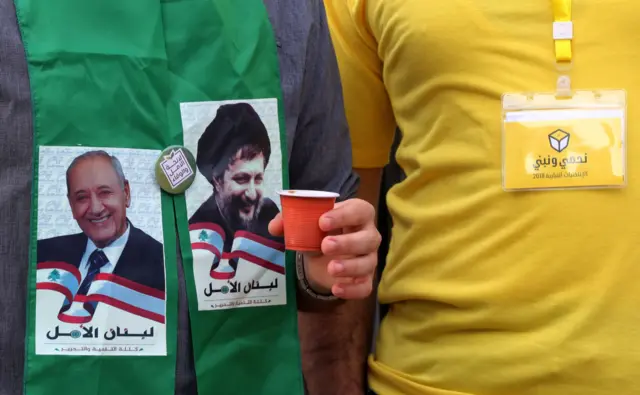 إلى اليمين رجل بقميص أصفر يرتدي شعار حزب الله الانتخابي نحمي ونبني وإلى اليسار ناخب يرتدي قميصاً أخضر وشعار حركة أمل الانتخابي لبنان الأمل مع صورة موسى الصدر وصورة نبيه بري ويحمل فنجان قهوة بلاستيكي