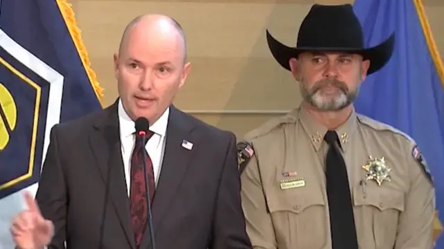 حاكم يوتا، سبنسر كوكس، يتحدث أمام ميكروفون مرتدياً بدلة رمادية وربطة عنق حمراء داكنة. يقف إلى جانبه شريف مقاطعة يوتا مايك سميث مرتدياً قبعة عريضة وقميصاً بنياً مزيناً بشارة شرطة على الجهة اليسرى من الصدر وبشارات "شريف" على كلا الكتفين.