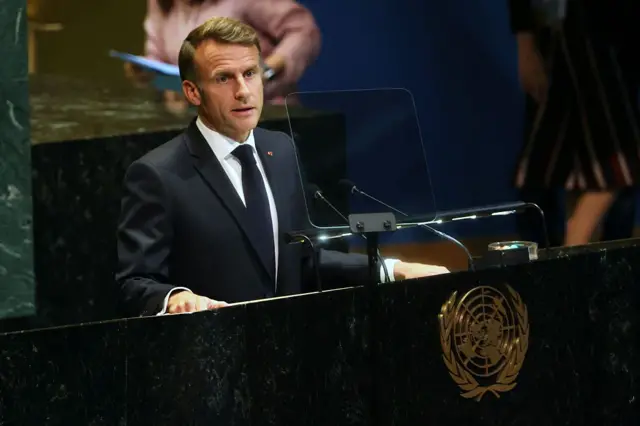 يرتدي ماكرون بدلة رسمية سوداء وقميصاً أبيض وربطة عنق سوداء، يقف على منصة الأمم المتحدة متحدثاً.