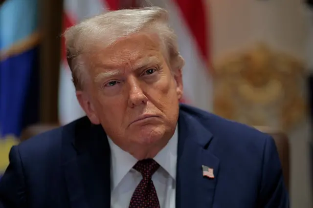 ترامب مرتدياً بدلة رسمية كحلية اللون وقميص أبيض وربطة عنق كحلية منقطة بالأحمر، ودبوس بالعلم الأمريكي. نظرات حادة تبرز من وجهه العابس في تلك اللحظة.