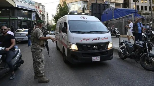 تسير سيارة إسعاف على طول شارع مزدحم متجاوزة رجلاً يرتدي زياً عسكرياً ويحمل بندقية هجومية