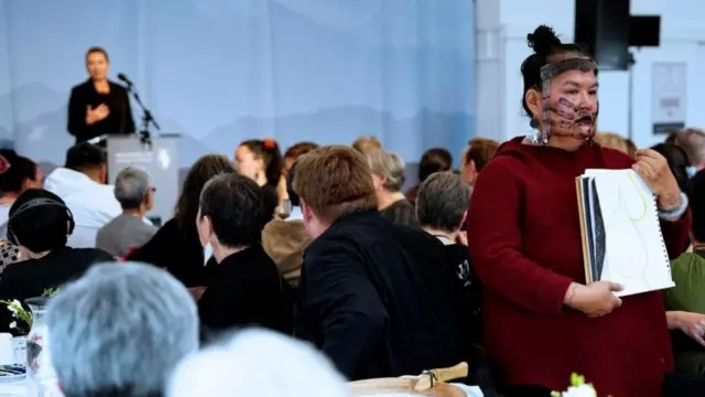 سيدة من السكان الأصليين بملابس نبيذية محاطة بحضور يجلسون على كراسٍ، تقف احتجاجا على كلمة رئيسة الوزراء الدانماركية خلال تقديم اعتذارها عن فضيحة تحديد النسل القسري في غرينلاند. وتحمل السيدة رسماً لبطن امرأة حامل. 