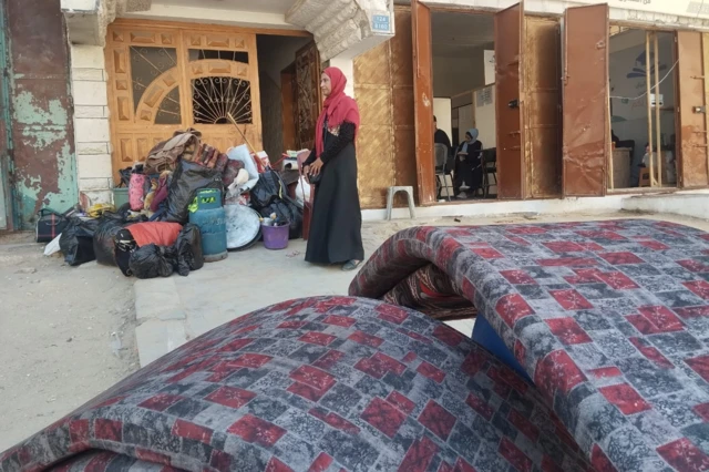 نوارة، امرأة أربعينية من غزة تقف أمام كومة من الأدباش والأغطية المحزومة بجوار قنينة غاز ومواعين وبراميل مياه، وأربعة أفرشة، تنتظر سيارة تنقلها هي وأطفالها الخمسة وأمتعتهم إلى جنوب القطاع. 