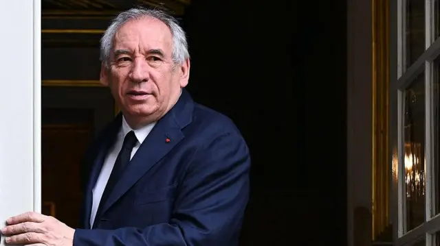 بايرو مرتدياً بدلة كحلية رسمية وقميصاً أبيض وربطة عنق كحلية. يقف بشكل جانبي ويضع يده على حافو حائط مقابل باب مفتوح. كبير في السن، بشرته بيضاء اللون وشعره أبيض، وعيناه لونهما أسود.