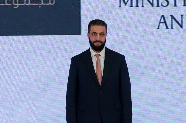 أحمد الشرع يرتدي بدلة رسمية كحلية وقميصاً أبيض وربطة عنق وردية اللون، شعره أسود ولديه لحية سوداء طويلة قليلاً.