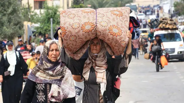 امرأتان محجبتان تسيران على طريق، هاربتان من منطقة رفح في 28 مايو/أيار 2024. إحداهما تحمل فراشاً كبيراً ملفوفاً على رأسها. خلفهما المزيد من الناس يسيرون ويحملون أمتعتهم، وسيارات محملة بالأمتعة، ورجل وطفل على دراجة هوائية محملة بحمولة ثقيلة.