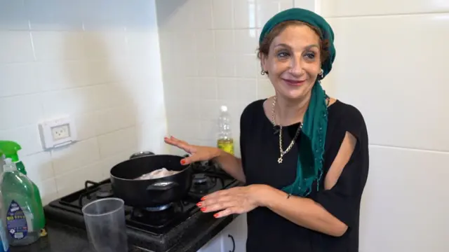 تقف امرأة بجانب قدح أسود مفتوح على الموقد. ترتدي وشاحًا أخضر حول رأسها، وبلوزة سوداء، وقلادة ذهبية عليها نجمة وهناك زجاجة زيت خلفها والجدران مغطاة بالبلاط الأبيض.