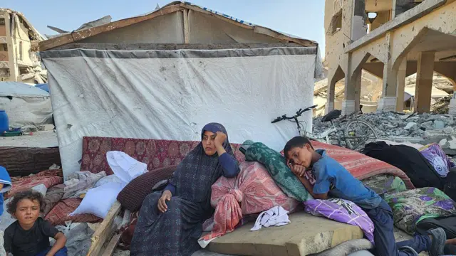 امرأة تجلس بين طفلين، أحدهما في سن ما قبل المدرسة، وصبي أكبر. يستريحان على كومة من المراتب والأمتعة أمام مأوى مؤقت مصنوع من القماش وقطع من الحشيش. يتكئ الصبي الأكبر على وسادة وكأنه يحاول النوم، بينما تُسند المرأة رأسها على يدها بتعبير حزين ومُتعب. يمكن رؤية بقايا المباني المدمرة على جانبي المأوى. التُقطت في منطقة المواصي، 17 سبتمبر/أيلول 2025.