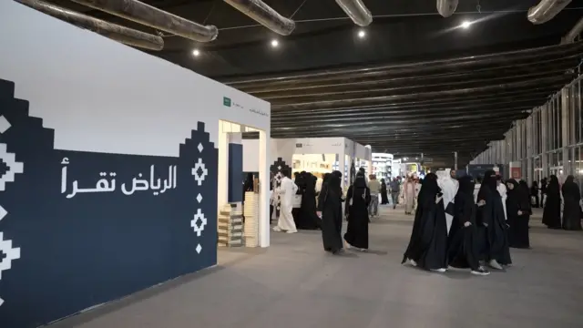 صورة تظهر نساء وفتيات ترتدي عباءات سوداء وتسير في أحد أروقة معرض الرياض الدولي للكتاب، ويسير كذلك رجال يرتدون جلاليب بيضاء. وهناك جدار كُتب عليه "الرياض تقرأ".