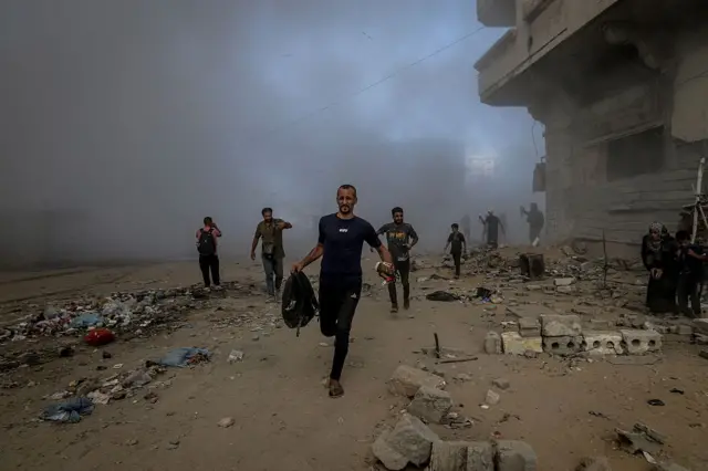 مجموعة من الأشخاص يحاولون النجاة من القصف الإسرائيلي، يركضون وسط أبنية مدمرة وداخن كثيف ناجم عن القصف الإسرائيلي على مدينة غزة.