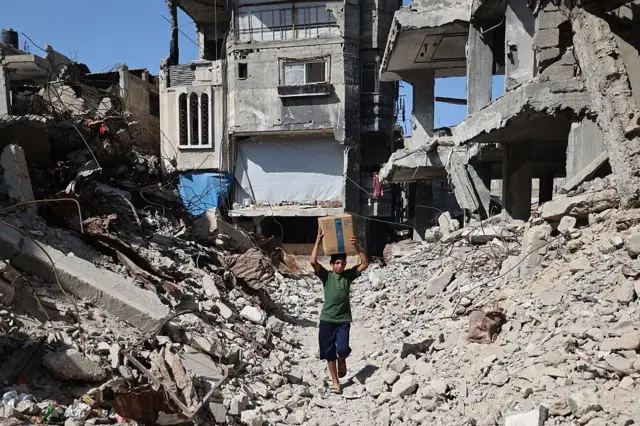 طفل فلسطيني مُشرّد يحمل صندوقًا من إمدادات الطوارئ التي قدّمها برنامج الغذاء العالمي بعد استلامه من نقطة توزيع مساعدات في مخيم النصيرات للاجئين وسط قطاع غزة، في 19 أكتوبر/تشرين الأول 2025.