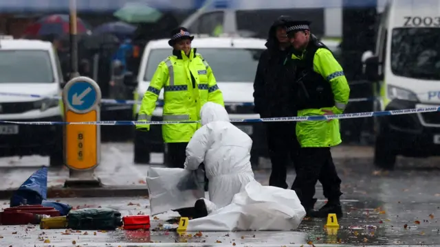 يقف ضابطان شرطيان يرتديان سترات عاكسة اللون وشخص آخر يرتدي معطفًا أسود بغطاء رأس في الشارع. يجلس شخص يرتدي زيًا أبيضًا جنائيًا على ركبته أمامهم. توجد سيارات شرطة في الخلفية ومعدات جنائية على الأرض.