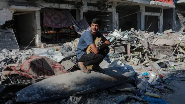 شاب يجلس على صاروخ إسرائيلي مُلقى على الأنقاض في وسط شارع بجوار منازل مُدمرة في مدينة غزة، قطاع غزة، 30 أكتوبر/تشرين الأول 2025، وسط وقف إطلاق نار بين إسرائيل وحماس. 