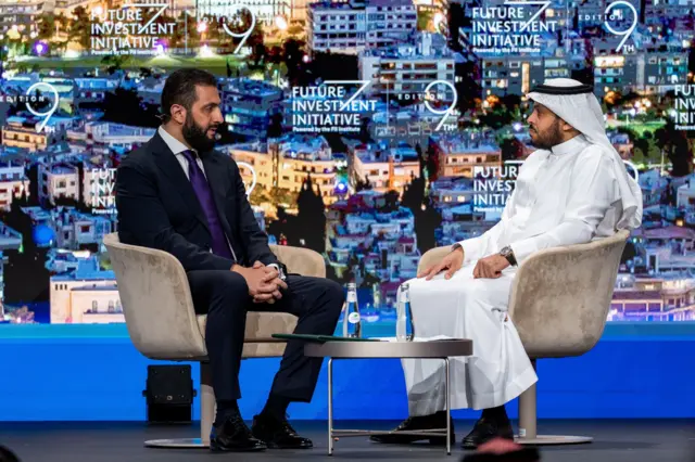 رئيس المرحلة الانتقالية في سوريا أحمد الشرع، خلال جلسة حوارية بالرياض يجلس على كرسي مرتدياً بدلة رسمية.