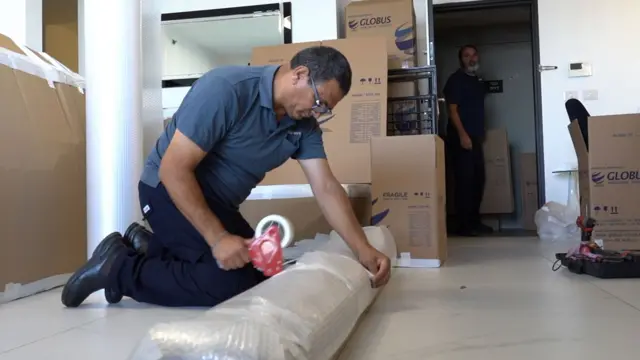 رجل يرتدي قميص بولو رمادي قصير وسروال داكن يركع على الأرض ويلصق شريطًا على غرض كبير ملفوف بفوّاز تبث. خلفه صناديق كرتونية ورجل آخر يراقب.