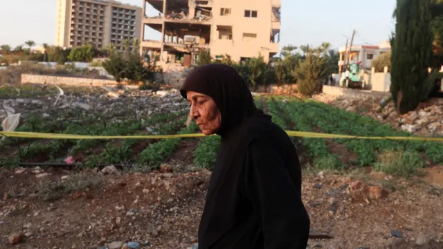 امرأة مسنة ترتدي حجابا اسود تسير بجانب حقل زراعي امام مبنى سكني مدمر جزئيا في ضوء شمس دافئة في مشهد يوحي بما بعد الدمار. خلفها مبان مهدمة وحطام وارض مزروعة وعلى يمينها شريط تحذير اصفر وتبدو في ملامحها حكمة السنوات وبقايا حزن.