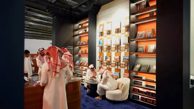 صورة تُظهر رجالاً يرتدون جلاليب بيضاء، بينما يرتدي بعضهم عقالاً، ويتصفحون بعض الكتب المعروضة في المعرض، وتحيط بهم أرفف وُضعت عليها كتب.