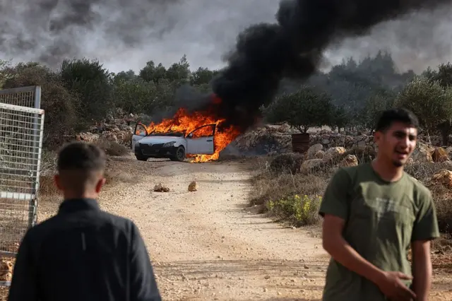 فلسطينيون يقفون بالقرب من سيارة محترقة أضرم فيها المستوطنون الإسرائيليون النار في محاولة لمنعهم من قطف الزيتون بالقرب من قرية ترمسعيا المحتلة بالضفة الغربية بالقرب من رام الله في 19 أكتوبر/تشرين الأول 2025.