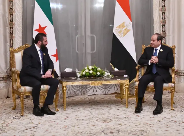 رئيس المرحلة الانتقالية في سوريا أحمد الشرع خلال لقائه الرئيس المصري عبدالفتاح السيسي يجلسان على كرسيين منفصلين وخلفهما العلمين السوري والمصري ويرتديان بدلة رسمية.