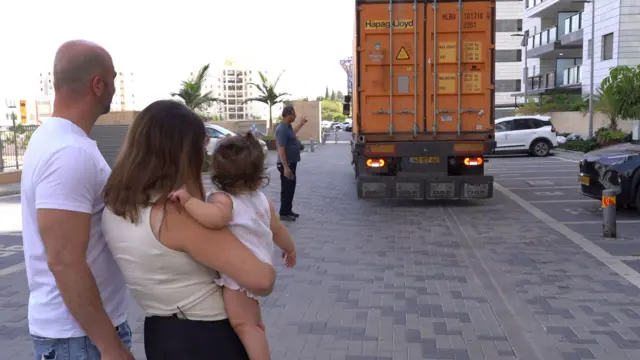 شاحنة نقل برتقالية تتراجع في شارع بينما يلاحظ نوفار وإيال اقترابها. يُرى الزوجان من الخلف - هو يرتدي قميصًا أبيض، وهي ترتدي قميصًا بدون أكمام بلون الكريم، وتحمل ابنتهما الصغيرة.