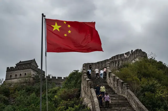 يُرى العلم الوطني على جزء من سور الصين العظيم بينما يتسلقه سياح خلال احتفالات اليوم الوطني والأسبوع الذهبي في هوايرو، على مشارف بكين. السماء ملبدة بالغيوم، الدرج الذي يتوسط السور الأثري طويل جداً، وتحيط الأشجار بطرفي السور.