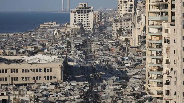 صورة من أعلى جزء من مدينة غزة، تُظهر منطقة واسعة مكتظة بالخيام على جانبي طريق موازٍ للبحر، مع مبانٍ متضررة على كلا الجانبين. تغطي الخيام كل الأراضي المفتوحة تقريبًا، باستثناء شريط ضيق حيث يقع الطريق.
