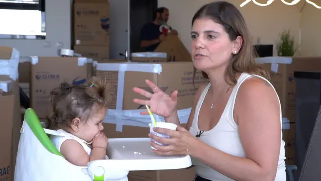 امرأة ذات شعر بني يصل إلى الكتف، ترتدي قميصاً صدرية باللون الكريمي، تطعم طفلاً صغيراً يجلس على كرسي مرتفع وهناك العديد من صناديق الكرتون في الخلفية.