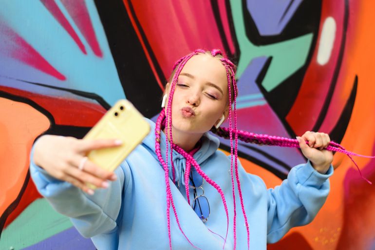 Caucasian teenage hipster girl with pink braids is using a smartphone against the background of a multicolored street wall.Summer concept.Generation Z style.Social media concept.; Shutterstock ID 2280791507; purchase_order: d; job: ; client: ; other: