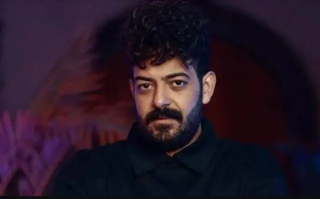 الليثي شعره أسود ولحية سوداء قصيرة، يرتدي قميصاً أسود ووراءه خلفية بتدرجات اللون الخمري والبنفسجي.