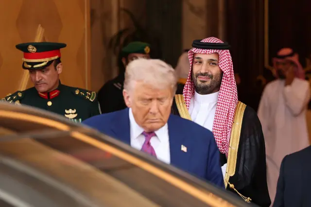 ترامب يقف مقابل سيارة يرتدي لباساً رسمياً كحلي اللون وربطة عنق تبدو خمرية اللون وقميصاً أبيض، خلفه تماماً ولي العهد السعودي مرتدياً اللباس التقليدي "العباءة السوداء والثوب الأبيض".