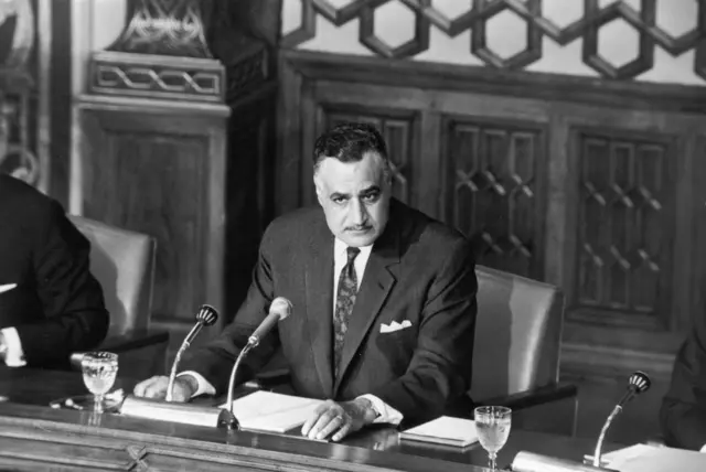 صورة للرئيس المصري الراحل جمال عبد الناصر جالساً، و يرتدي سترة وقيمصاً و ربطة عنق وأمامه ميكروفون.