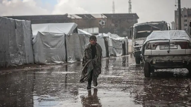 طفل فلسطيني يركض تحت المطر في غزة بينما تظهر خيام النازحين من خلفه وقد غمرتها المياه