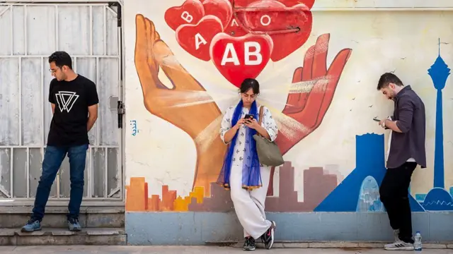 شابة ترتدي بنطالاً وقميصاً يغلب عليهما اللون الأبيض، القميص مزركش باللون الأزرق وعلى كتفيها شال بلون أزرق غامق. خلفها رسم على جدار يظهر أنواع فصائل الدم وفق تصنيف ABO، داخل رسوم قلب حب. الشابة تقف في المنتصف بين شابين.