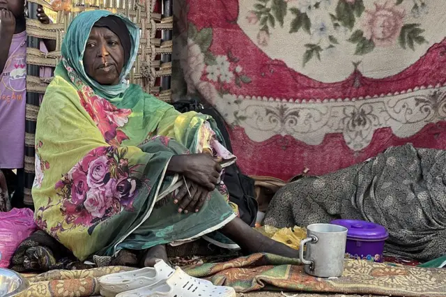 سيدة سوادانية نازحة تفترش الأرض وترتدي الزي السوادني التقليدي الثوب، وبجوارها نعلين.