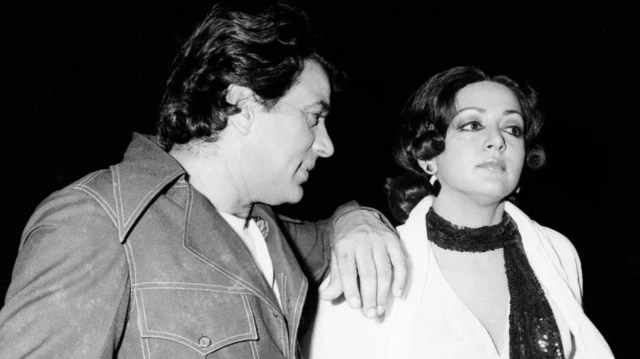 دارمندرا واضعاً يده على كتف زوجته الممثلة الهندية هيما ماليني، يرتدي كلاهما ملابس أنيقة في صورة بالأبيض والأسود.