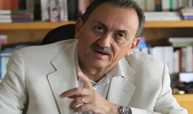 سيمون كرم يرتدي بدلة رسمية بيضاء اللون وخلفه مكتبة صغيرة