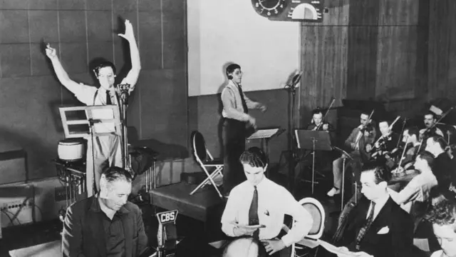 في استوديو إذاعة سي بي إس، يتدرب أورسون ويلز على أحد برامجه، واقفاً أمام الميكروفون مرتدياً قميصاً أبيض وربطة عنق داكنة، وذراعيه مرفوعتان بشكل درامي في منتصف العرض. على يمينه، يُجهّز أعضاء الأوركسترا آلاتهم الموسيقية، بينما يحمل الممثلون في المقدمة نصوصًا جاهزة لإشاراتهم.
