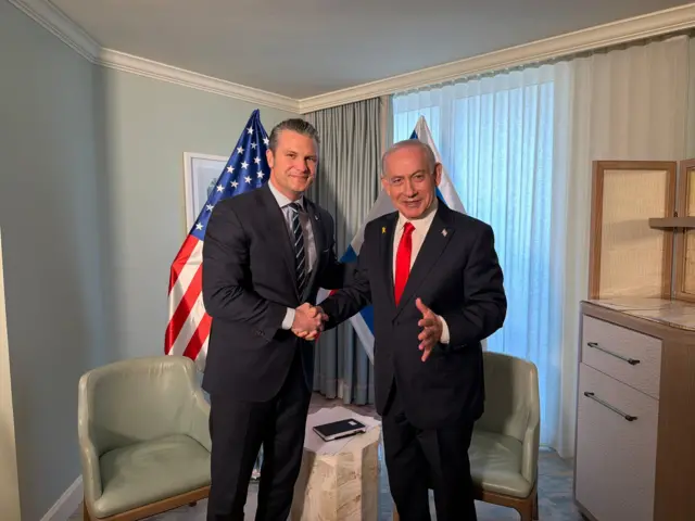 رئيس الوزراء الإسرائيلي بنيامين نتنياهو يلتقي وزير الحرب الأمريكي، بيت هيغسيث في فلوريدا. ويقف نتنياهو مصافحاً هيغسي، ويرتدي نتنياهو سترة سوداء وقميصاً أبيض وربطة عنق حمراء. بينما يرتدي هيغسيث قميصاً رصاصي اللووسترة سوداء وربطة عنق تمزج بين اللونين الأزرق والأبيض. وفي الخلفية علم الولايات المتحدة وإسرائيل.