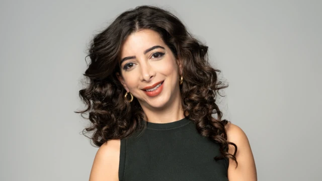 امرأة ذات شعر أسود مجعّد، ترتدي قميصاً أسود بلا أكمام وتضع أقراطاً ذهبية دائرية.