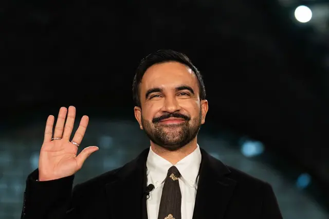 زهران ممداني يرفع يده للأعلى لحلف اليمين، مُبتسماً، ويرتدي سترة سوداء وربطة عنق بنية اللون وقميصاً أسود.