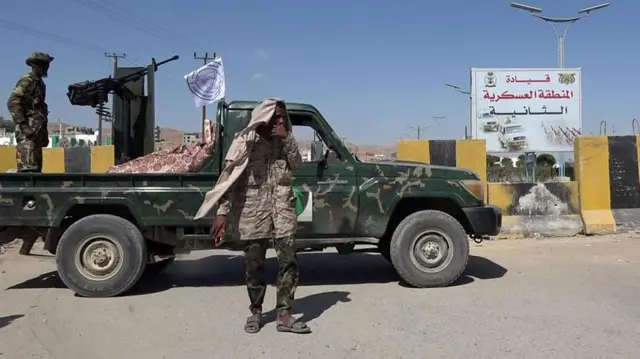 رجل بزي عسكري يضع سترة على رأسه وخلفه آلية عسكرية تحمل رشااشا آليا مع عسكري آخر واقف خلفها. في الخلف لافتة تحمل اسم قيادة المنطقة العسكرية الثانية