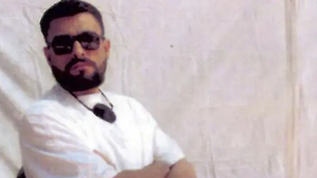 أبوزبيدة يرتدي نظارة شمس سوداء وتي شيرت أبيض وبنطلون أبيض ويجلس على كرسي